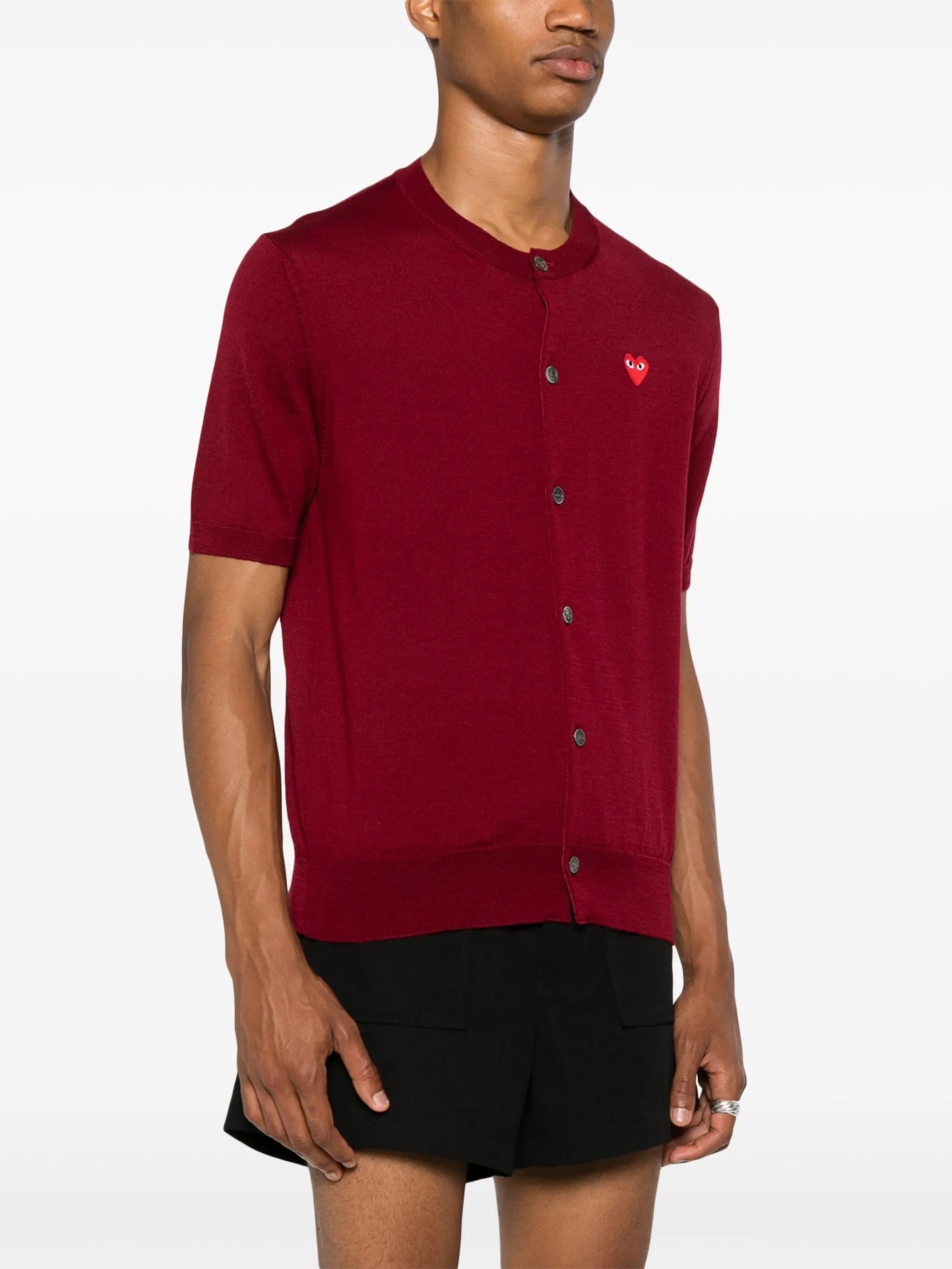 Side profile of male model wearing the red short-sleeve cardigan with a red heart logo from Comme des Garcons Play on a white background