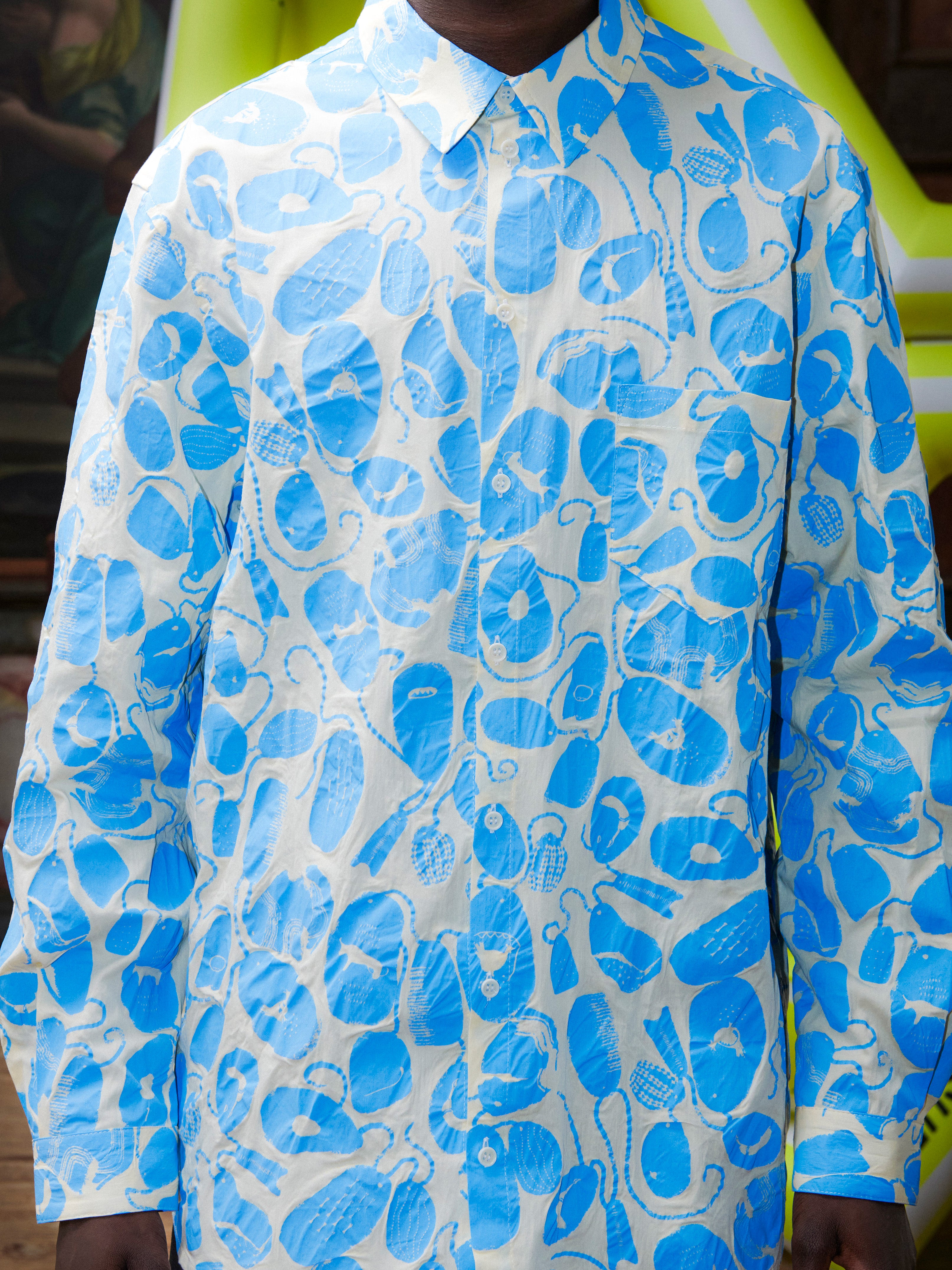 Close-up of model wearing the Henrik Vibskov New Blou Shirt in Ecru Amulets, front view, showing the off-white shirt with blue amulets pattern, collar, front buttons, and chest pocket.