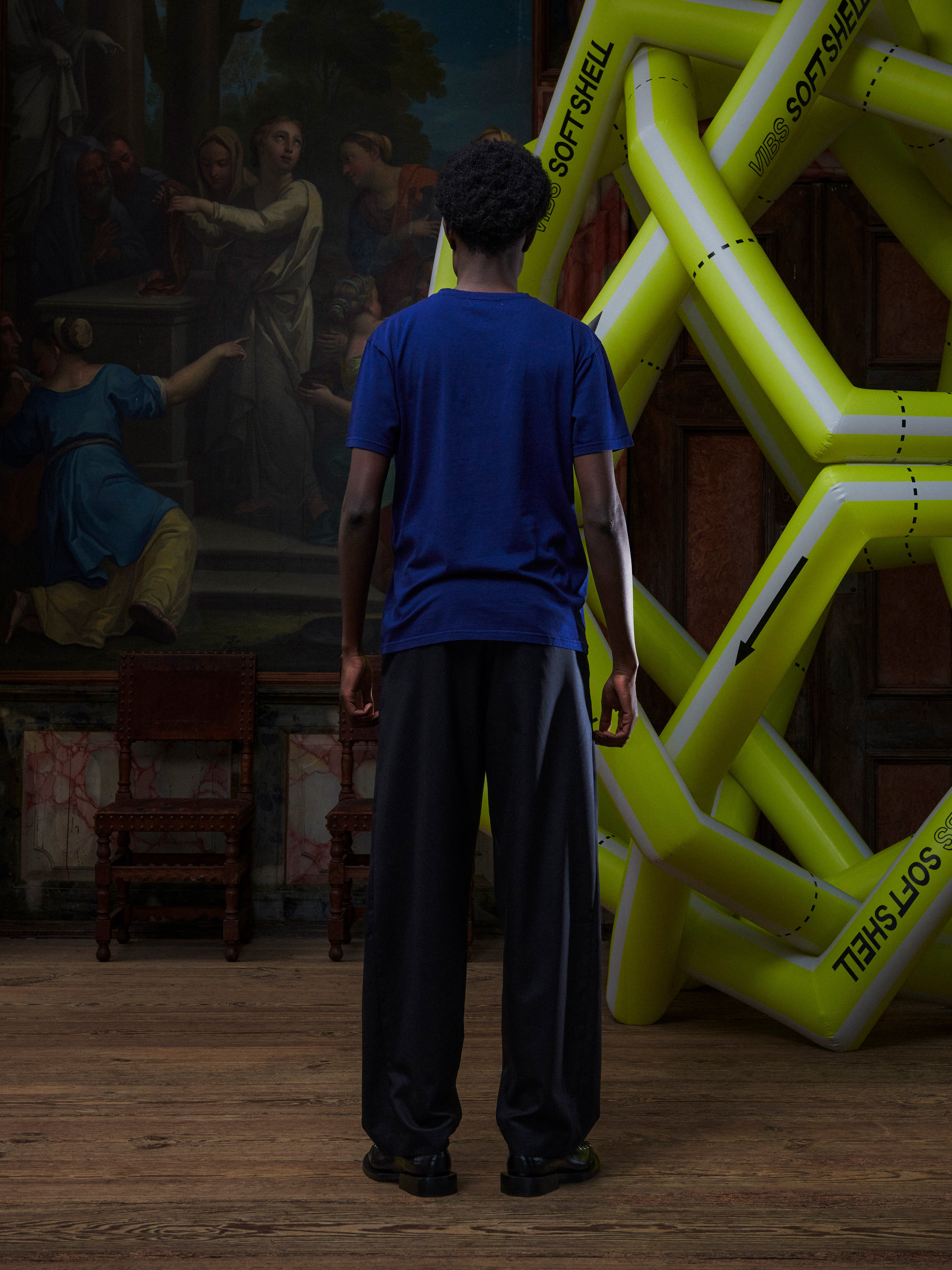 Back view of male model wearing the navy blue t-shirt with a pink rooster illustration by Henrik Vibskov in a setting with an inflatable structure