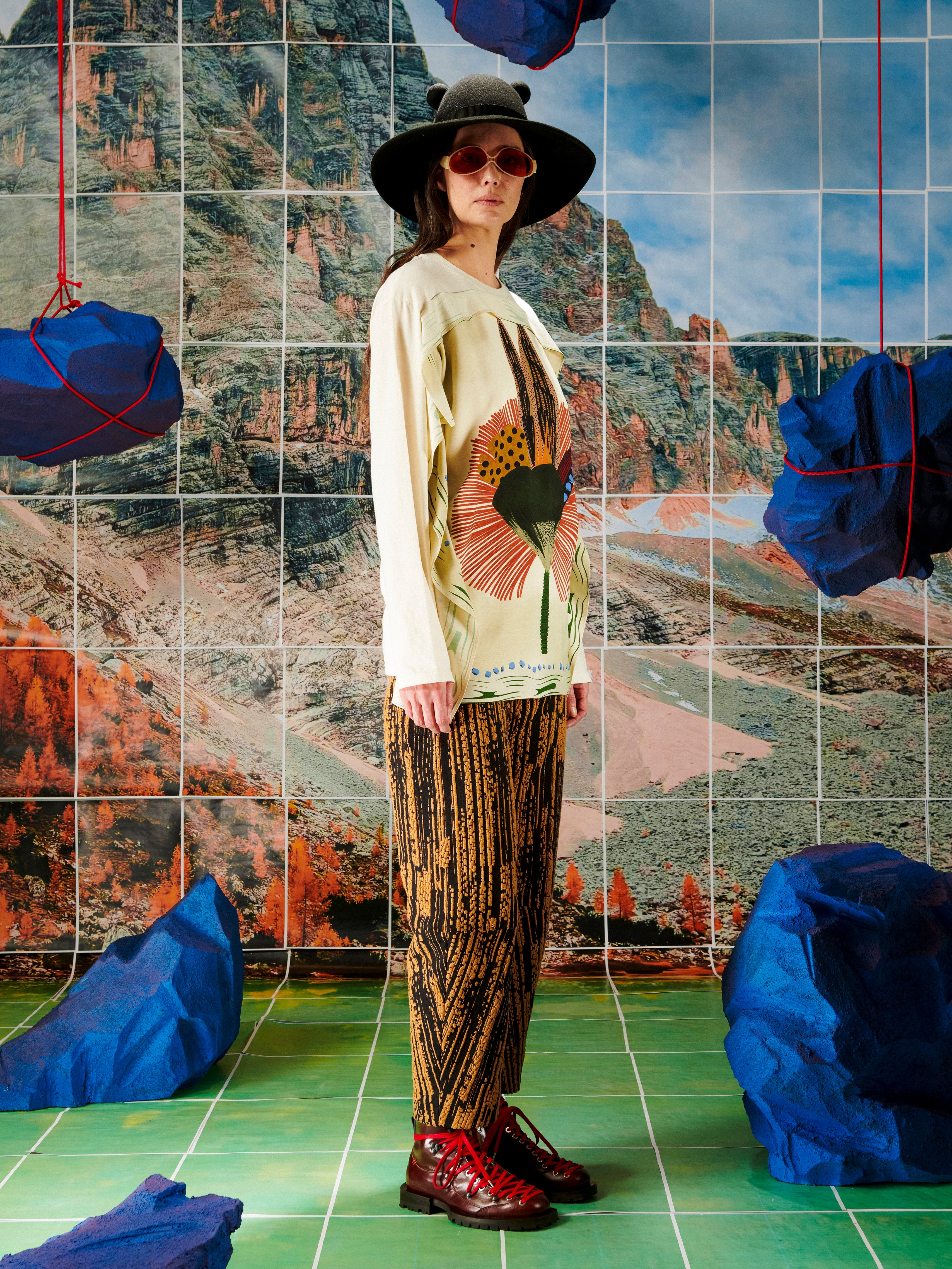 Person wearing a hat and sunglasses standing in front of a tiled wall with mountain and blue rocks designs.