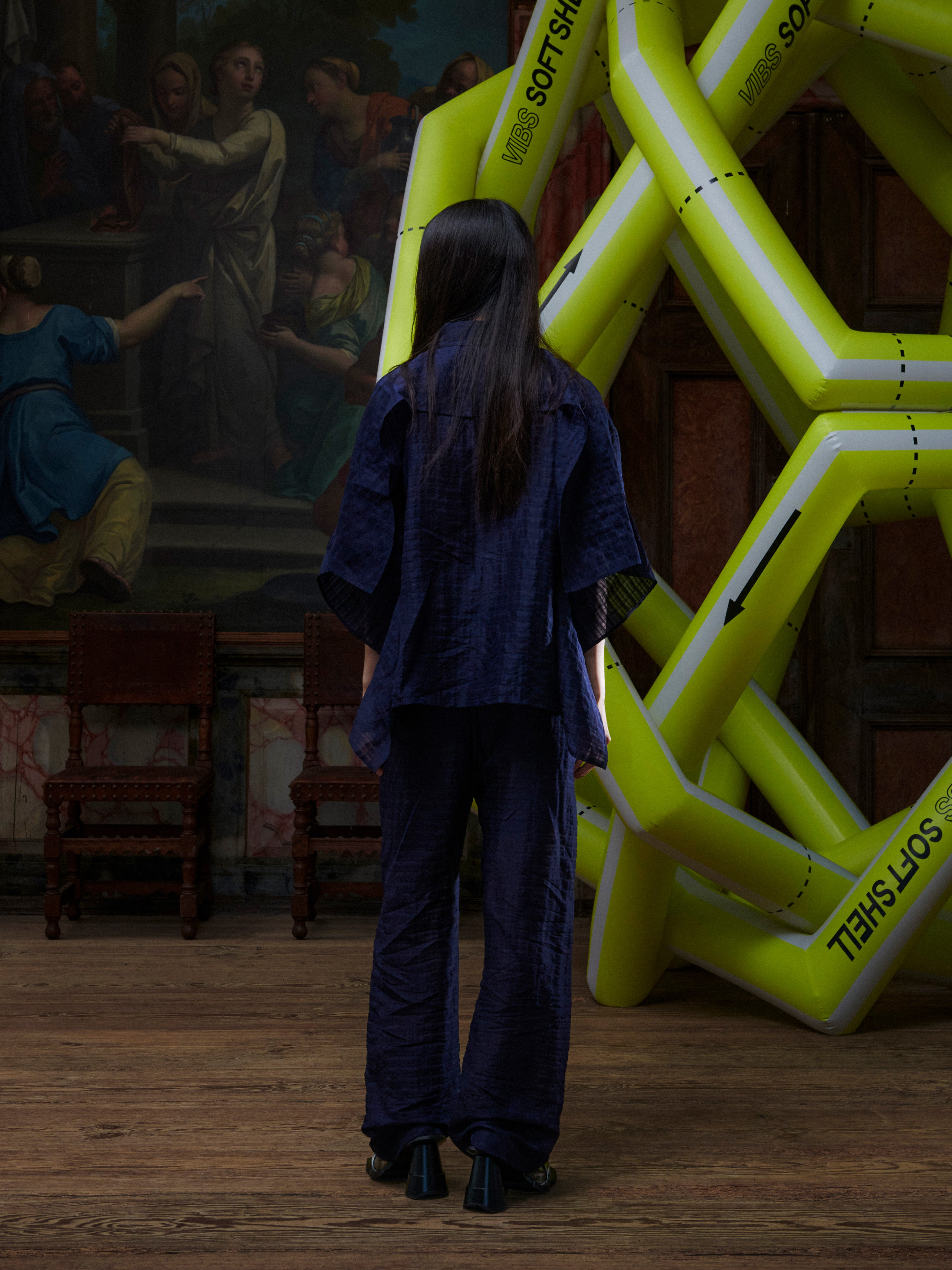 Person in a dark outfit standing in front of a classical painting with a large yellow inflatable structure.