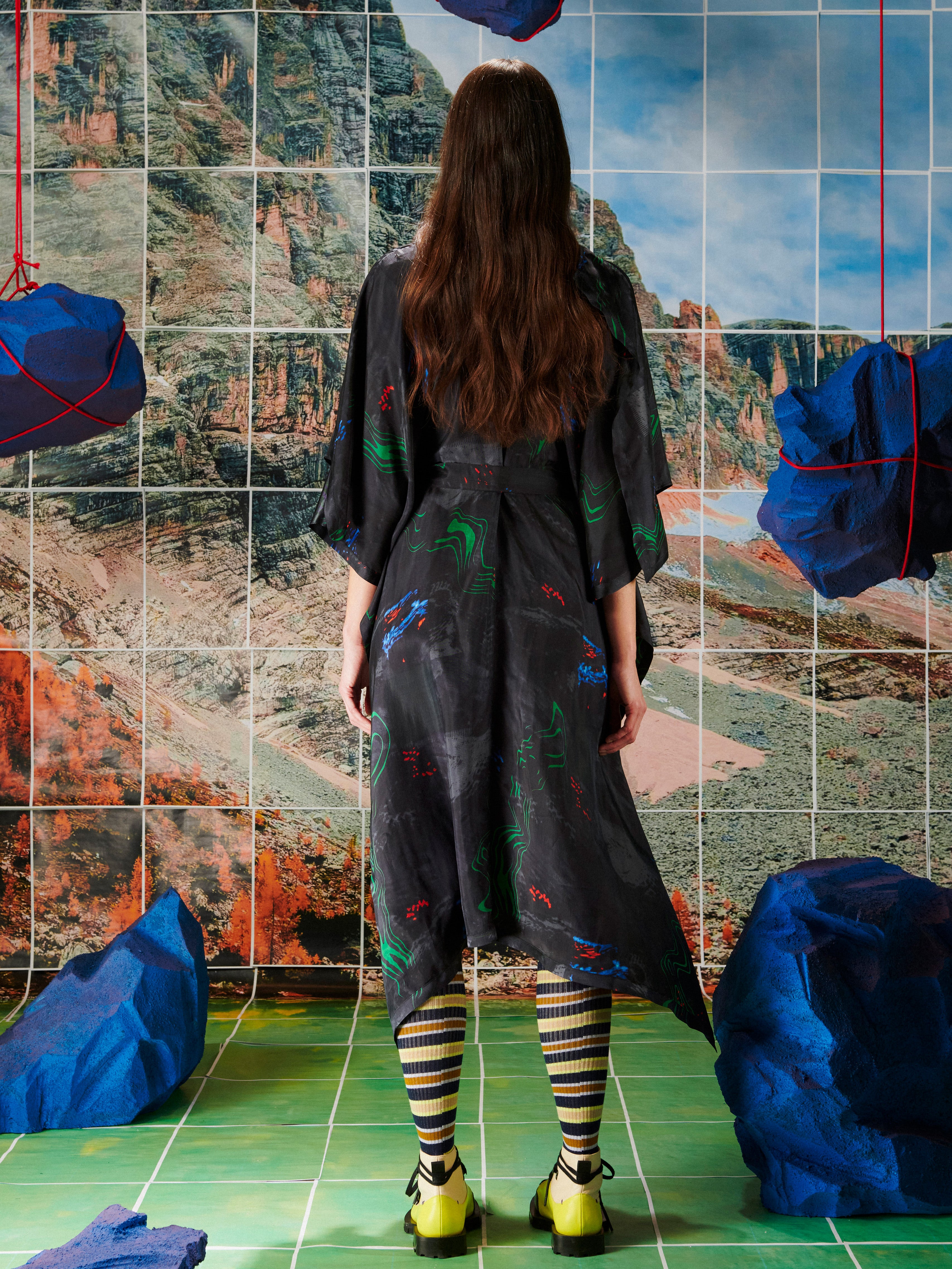 Woman in a black dress with colorful patterns from Henrik Vibskov standing in front of a tiled wall with mountain design.