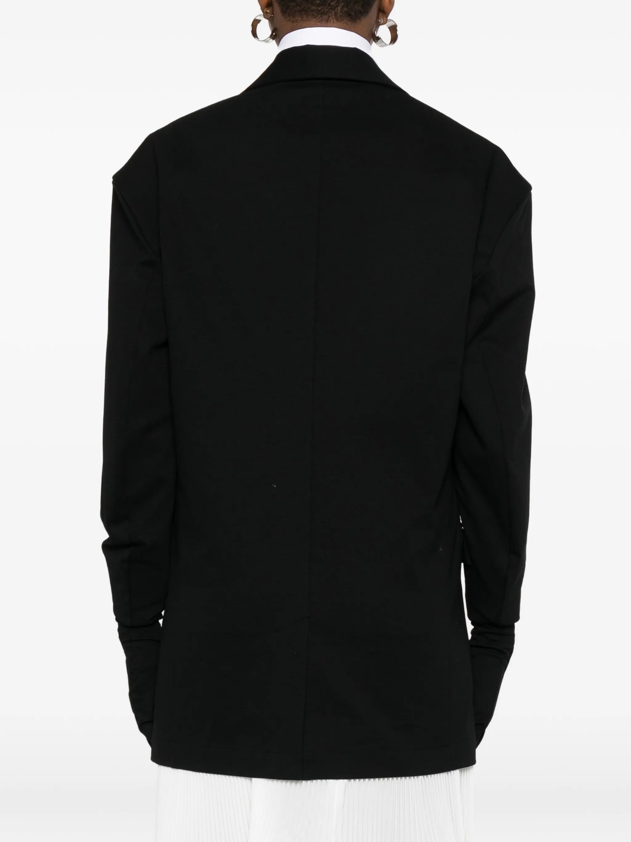 Back view of model wearing the black blazer with gloves attached to sleeves from Issey Miyake on a white background