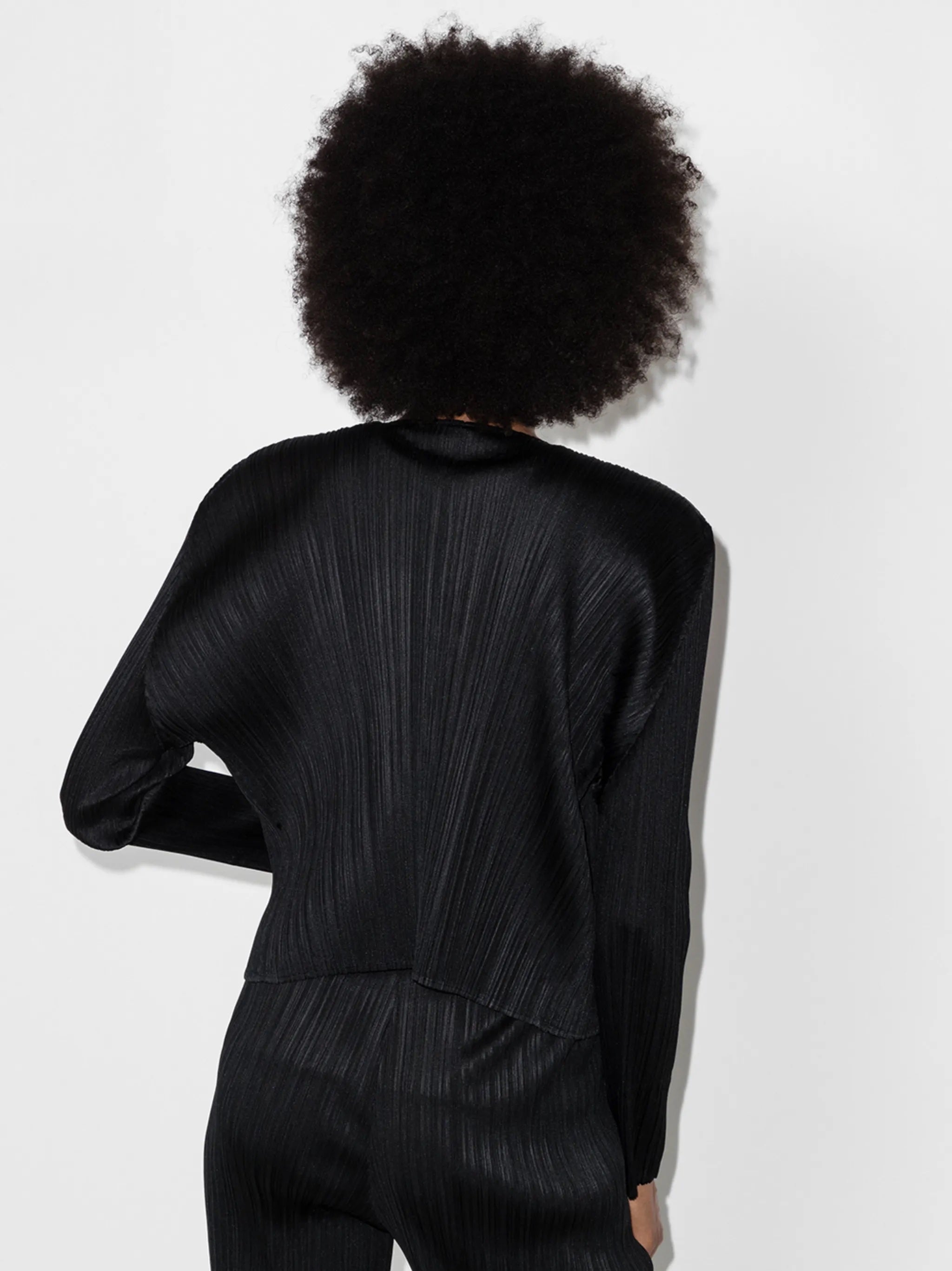 Person wearing a black pleated outfit from Pleats Please Issey Miyake on a white background