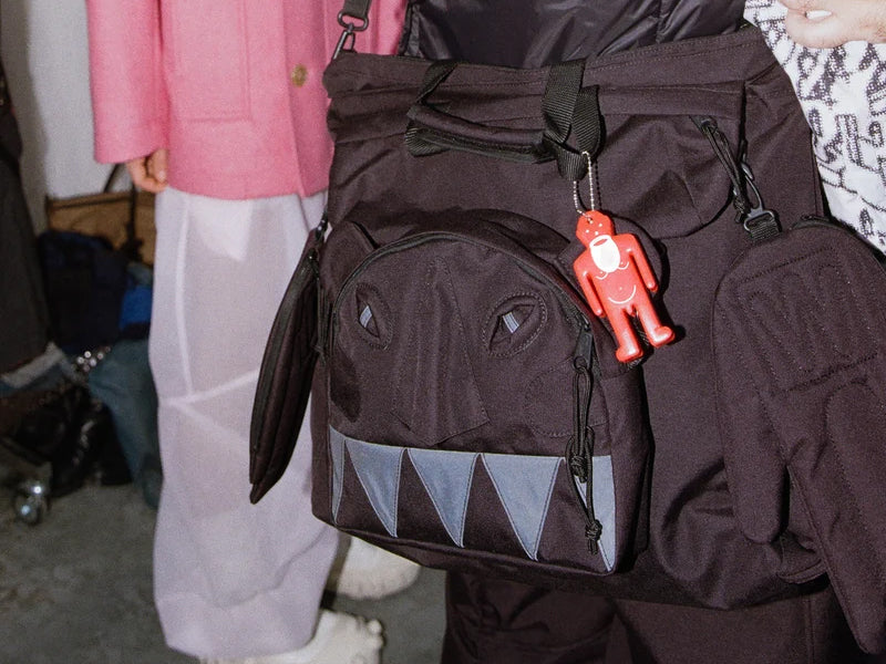 Walter Van Beirendonck Monster Carry Bag in Black, styled close-up showing sculptural monster face, triangular teeth motif, padded side hand details and red character charm.