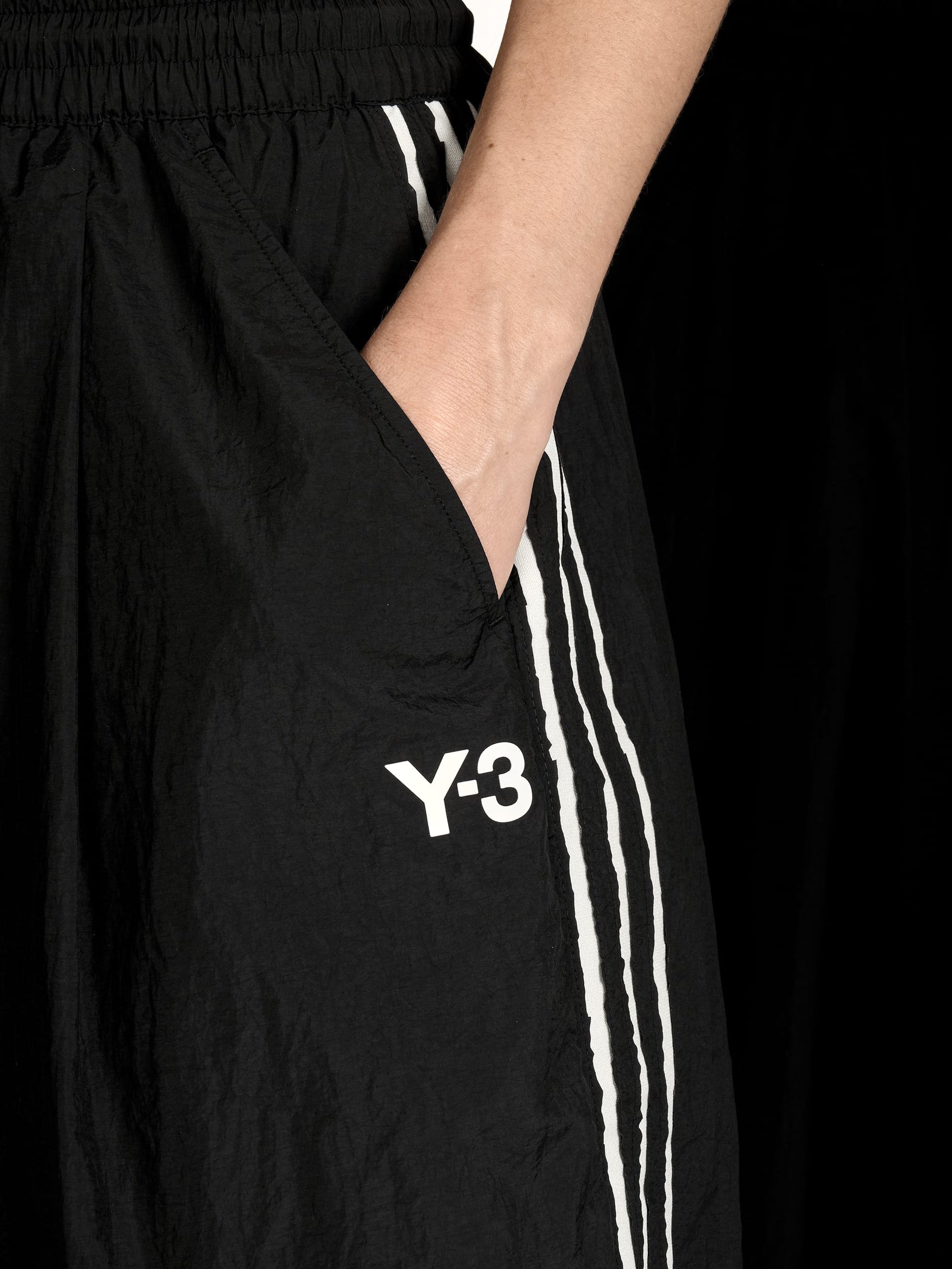 Close-up side-front of a female model wearing the Y-3 Raw Edge 3-Stripes Wide Tracksuit Bottoms in Black, highlighting the wide-leg drape, adjustable bungee cord hems, and angled side pocket details.