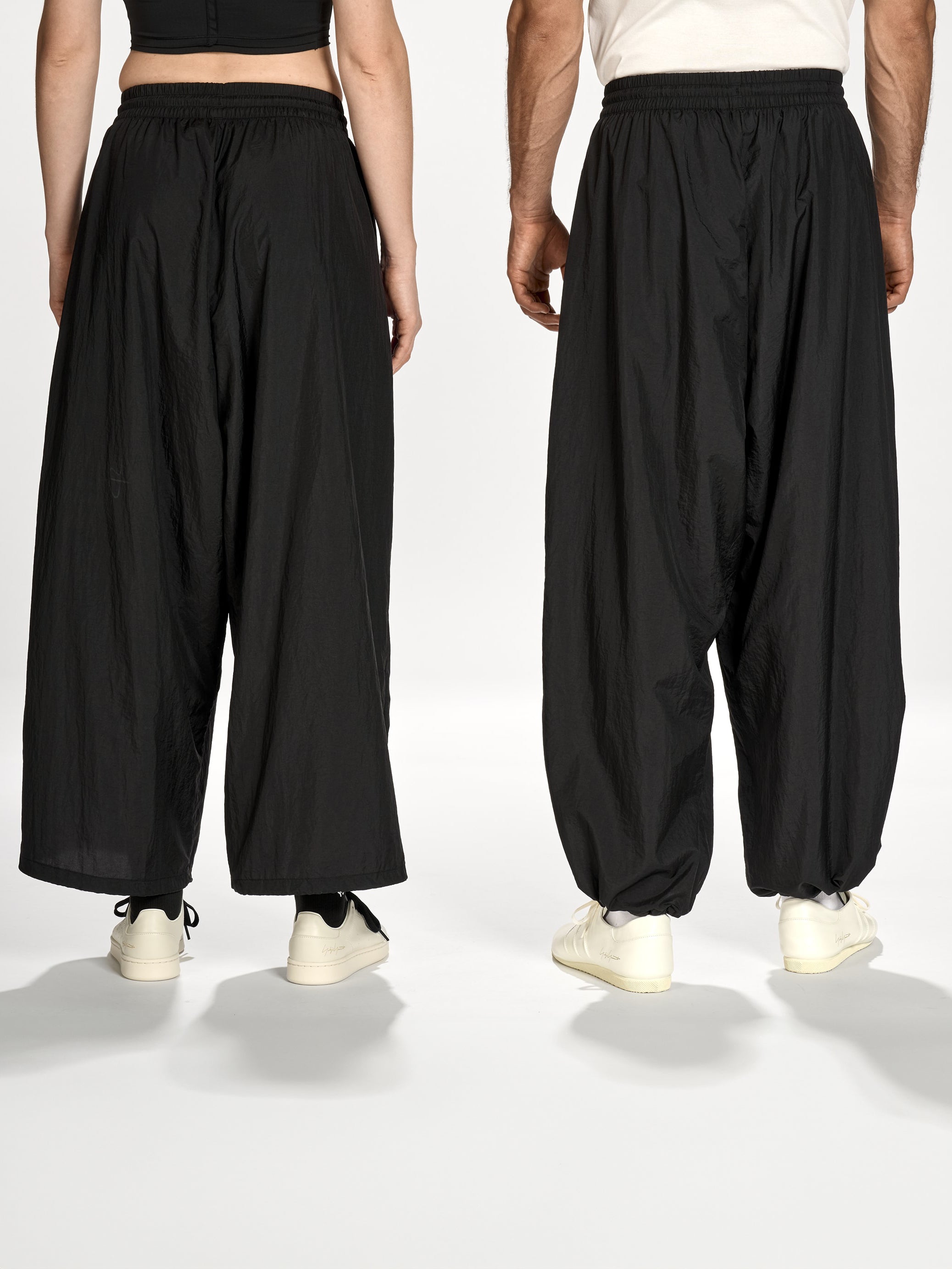 Back view of a female and male model wearing the Y-3 Raw Edge 3-Stripes Wide Tracksuit Bottoms in Black, highlighting the deconstructed raw-cut 3-Stripes, relaxed silhouette, and clean rear detailing.