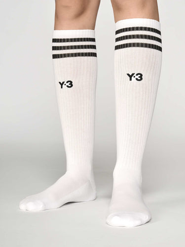 Front view of a model wearing the Y-3 Stripes High Socks in White, showing the knee-length fit, engineered 3-Stripes cuff, and embroidered Y-3 logo at the centre front.