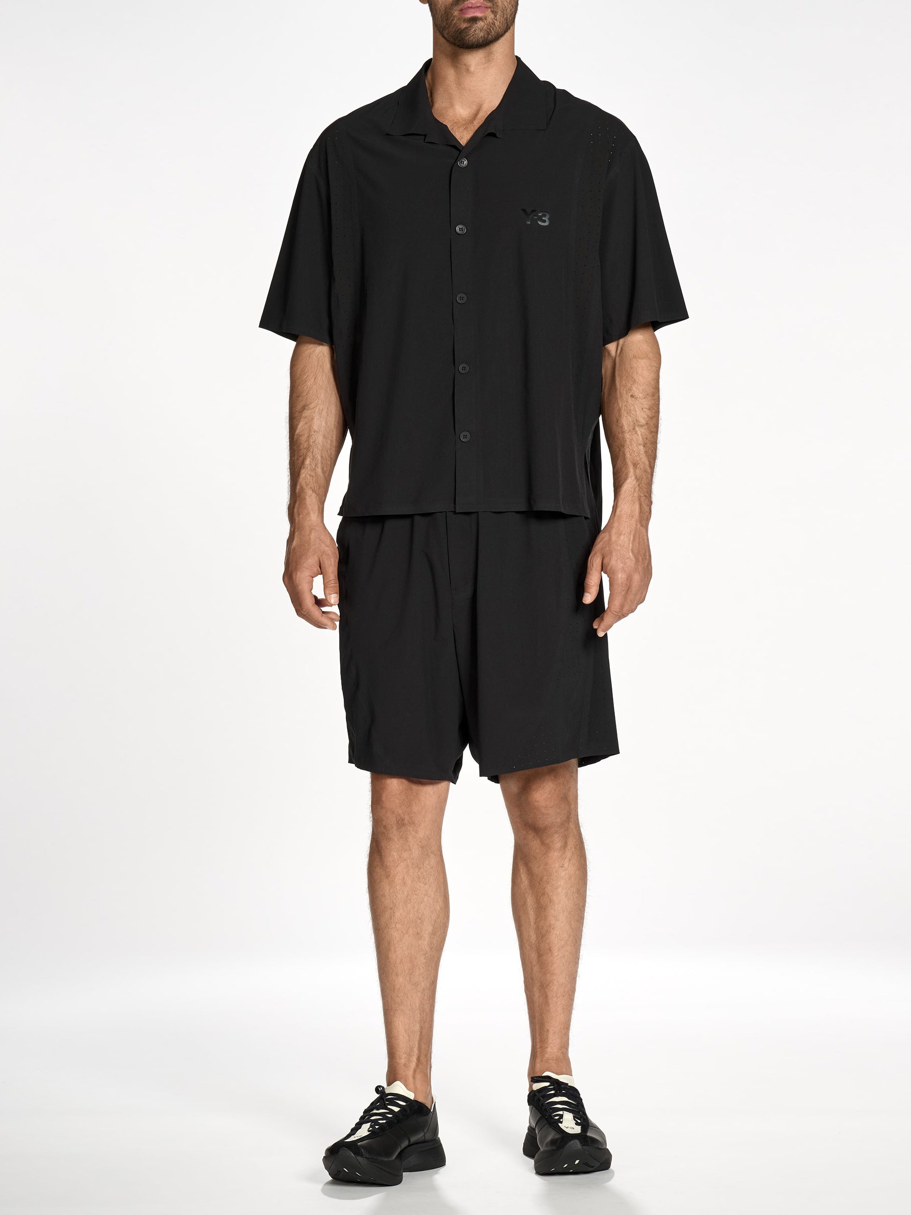 Front view of a male model wearing the Y-3 Ultralight Woven Shirt in Black, showcasing the sleek fit, short sleeves, asymmetrical bonded ventilation slits, and centre-front button closure.