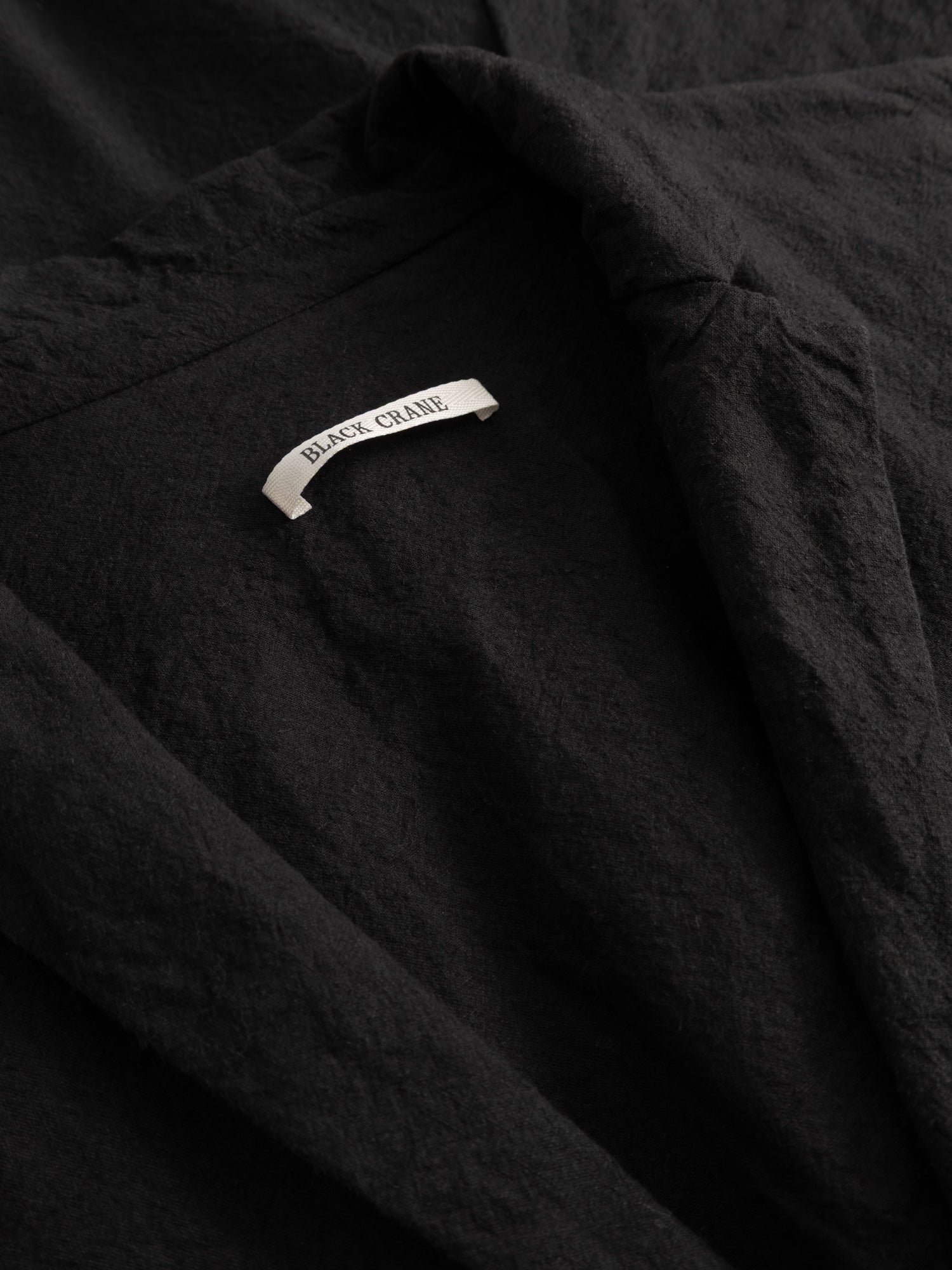 Close-up of the Black Crane Crinkled Coat showing the crinkled black fabric texture and the brand label at the neckline.
