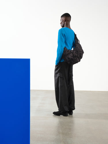 Male model wearing Côte&Ciel Hyco S Otra Gathered crossbody bag in black, styled across the body, highlighting its ergonomic sculptural shape and ruched nylon texture.