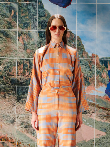 Person wearing an orange and blue checkered outfit with sunglasses, standing in front of a scenic mountain background.