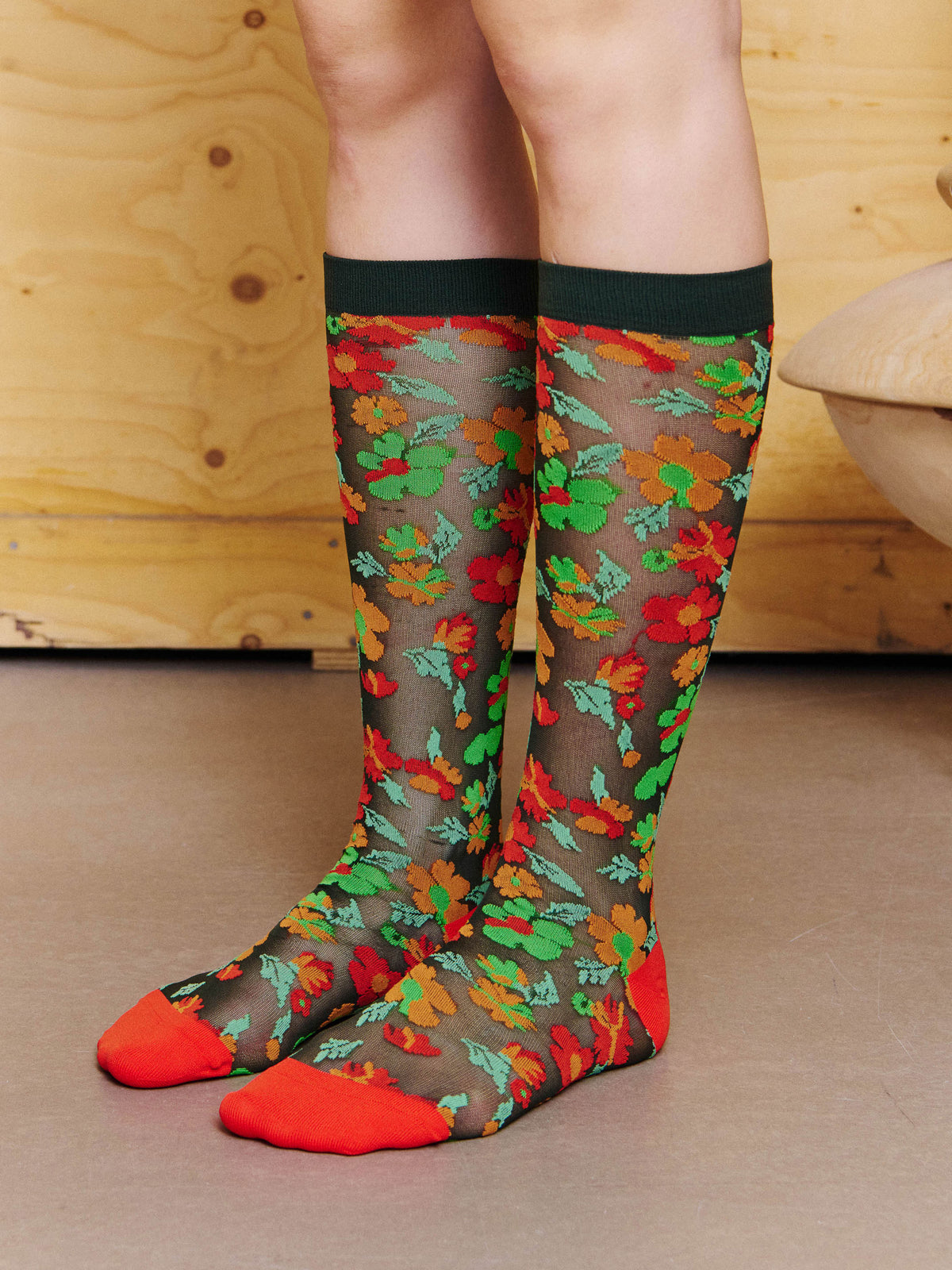 Angled view of model wearing the Henrik Vibskov Bouquet Socks in Orange Floral Affairs, showing the floral motif, semi-sheer knit and contrast toe and heel.