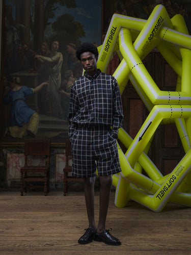 Front full-body view of a male model wearing the Claus Shorts in Black Checks from Henrik Vibskov Spring/Summer 2026 Studio Collection, highlighting the multicoloured check pattern, side pockets, and relaxed unisex fit.