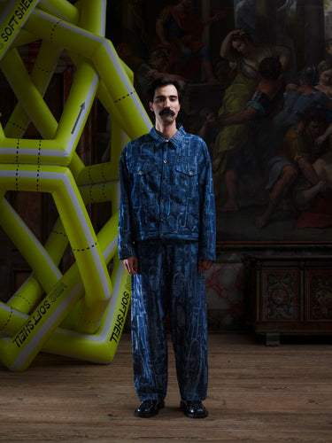 Male model wearing the Curved Denim Pants from the front, showing the straight-leg fit, grey-blue denim, and abstract instrument print. Model is 185cm tall and wears size M.