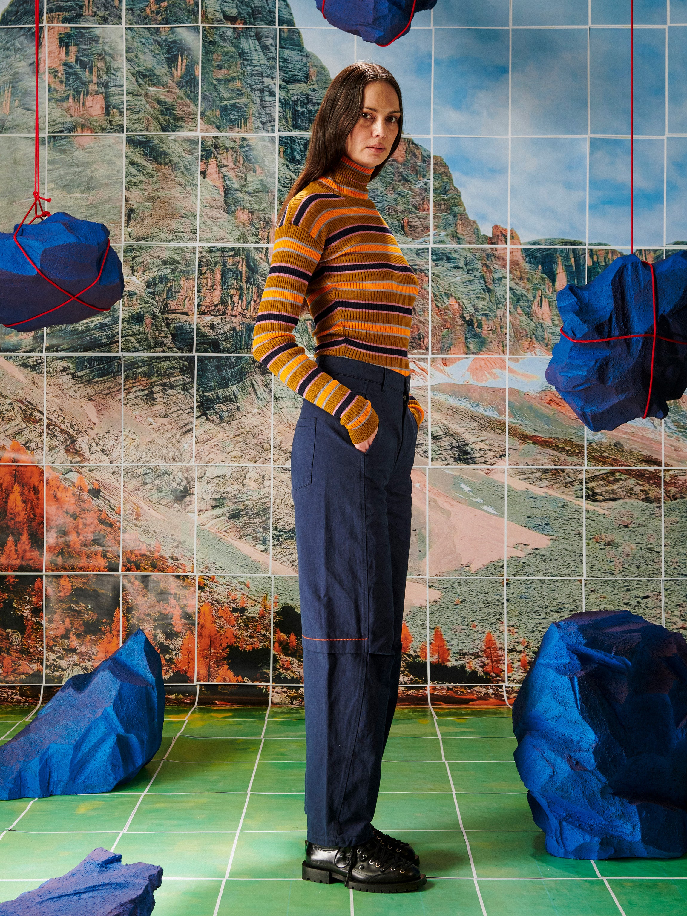 Woman in striped sweater and pants standing in front of a tiled wall with landscape designs.