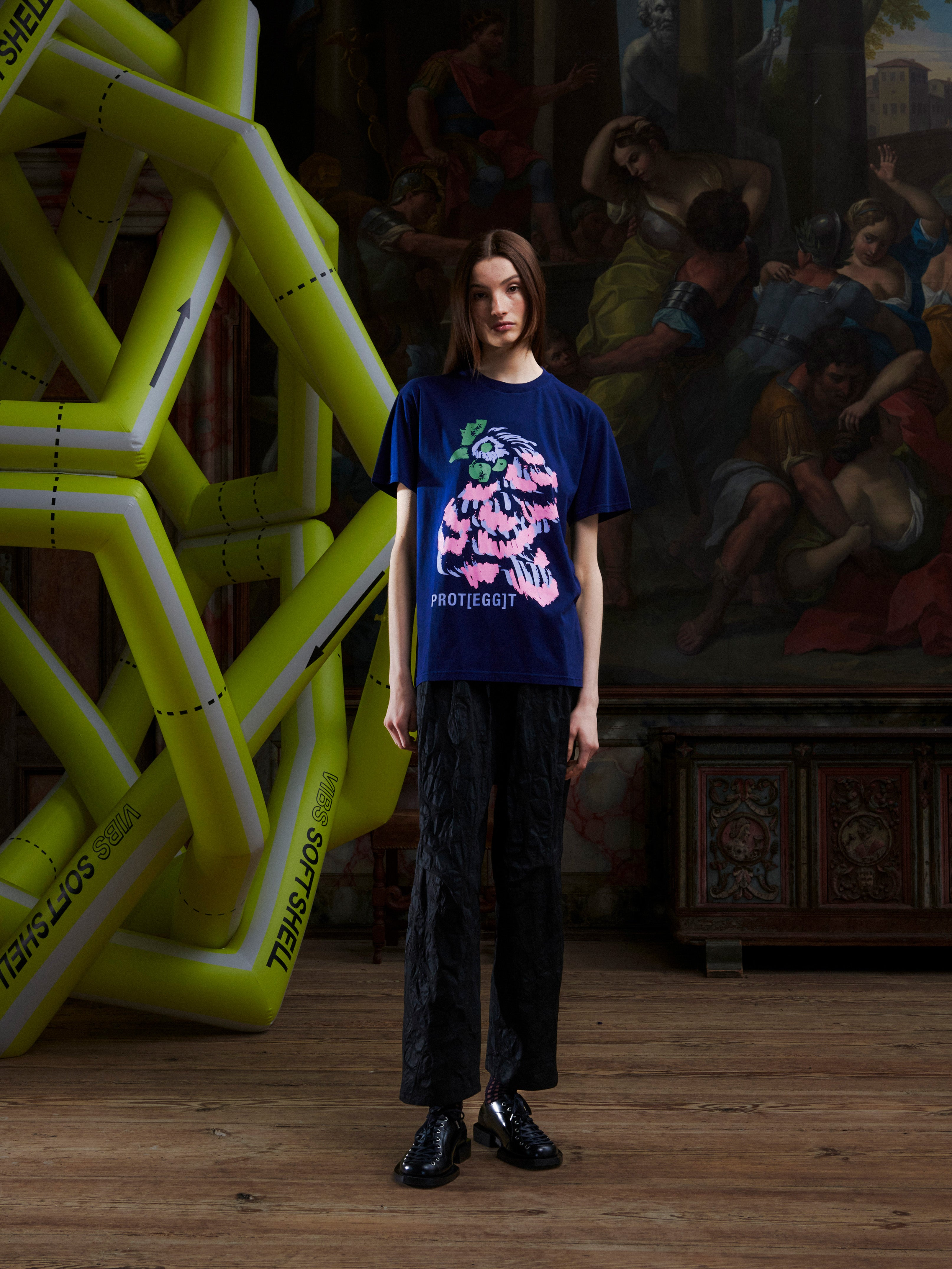 Female model wearing the navy blue t-shirt with a pink rooster illustration by Henrik Vibskov in a setting with an inflatable structure