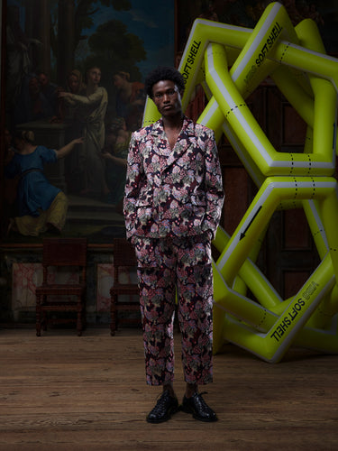 Male model wearing a patterned suit by Henrik Vibskov, standing in front of a large inflatable sculpture with a painting in the background.