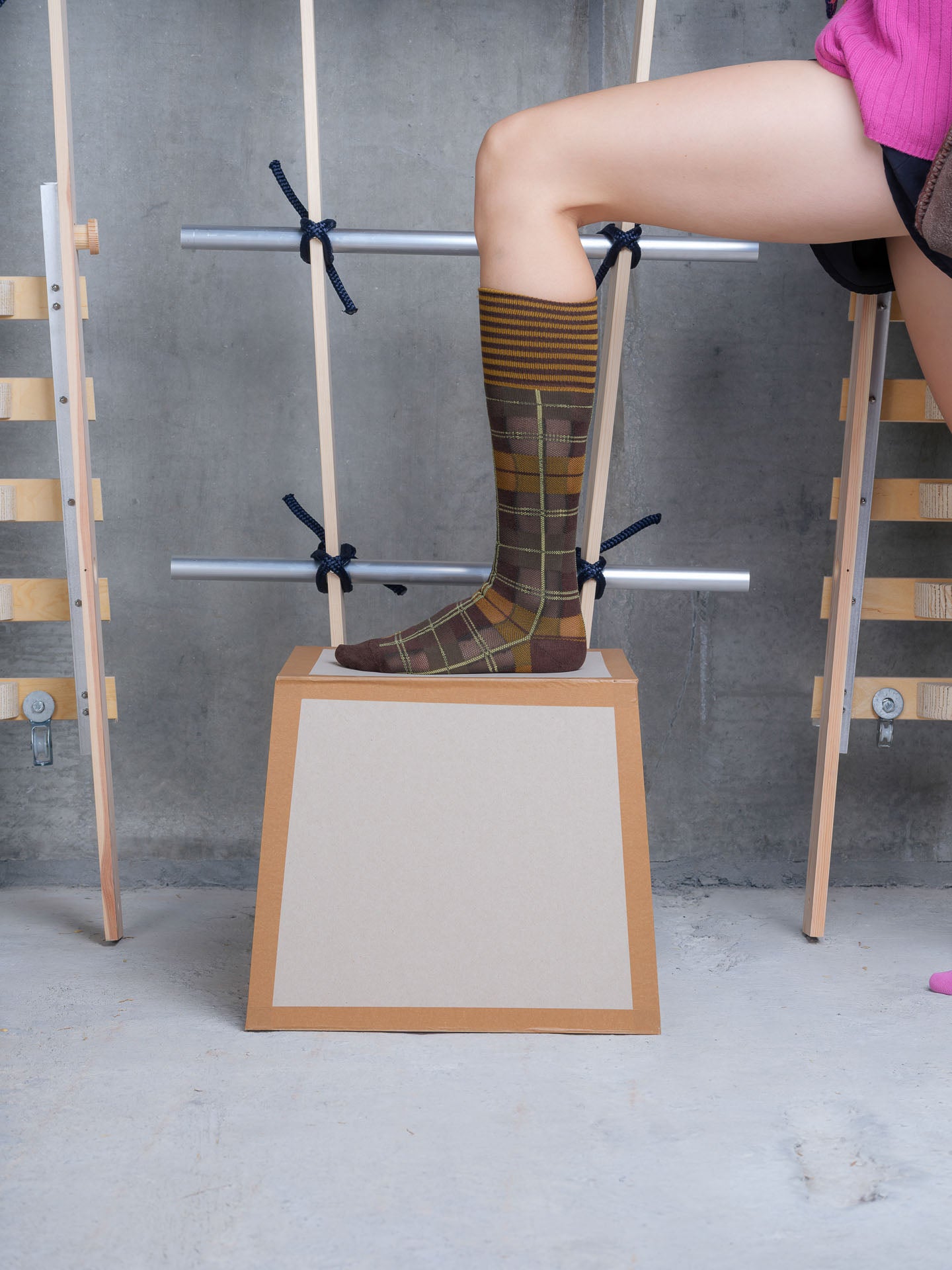 Model wearing Henrik Vibskov Ribbit Knee Sock in olive green and brown check with striped ribbed cuff and contrast heel and toe