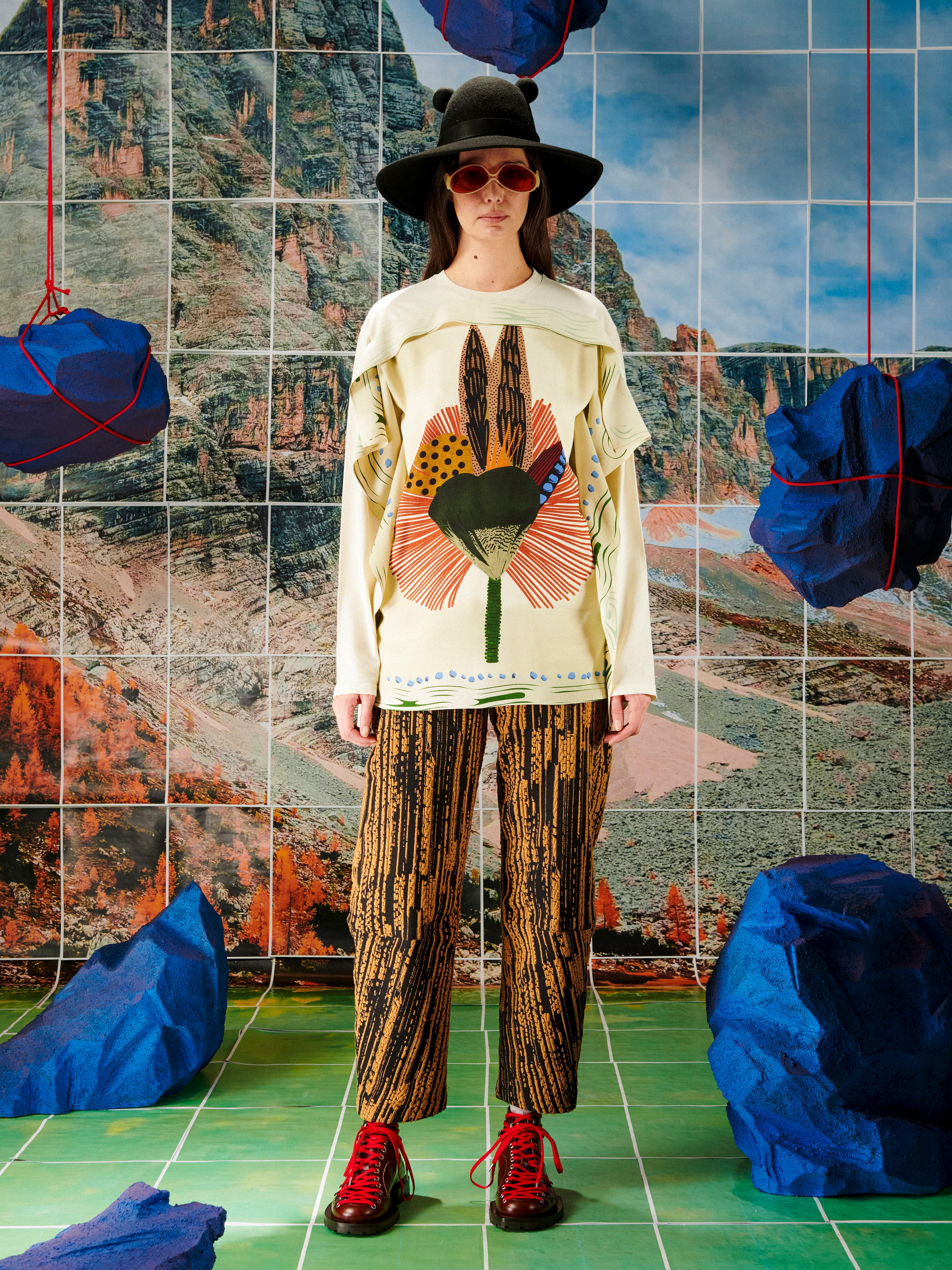Person wearing a patterned outfit with a colorful top and striped pants, standing in front of a tiled wall with a mountainous landscape.
