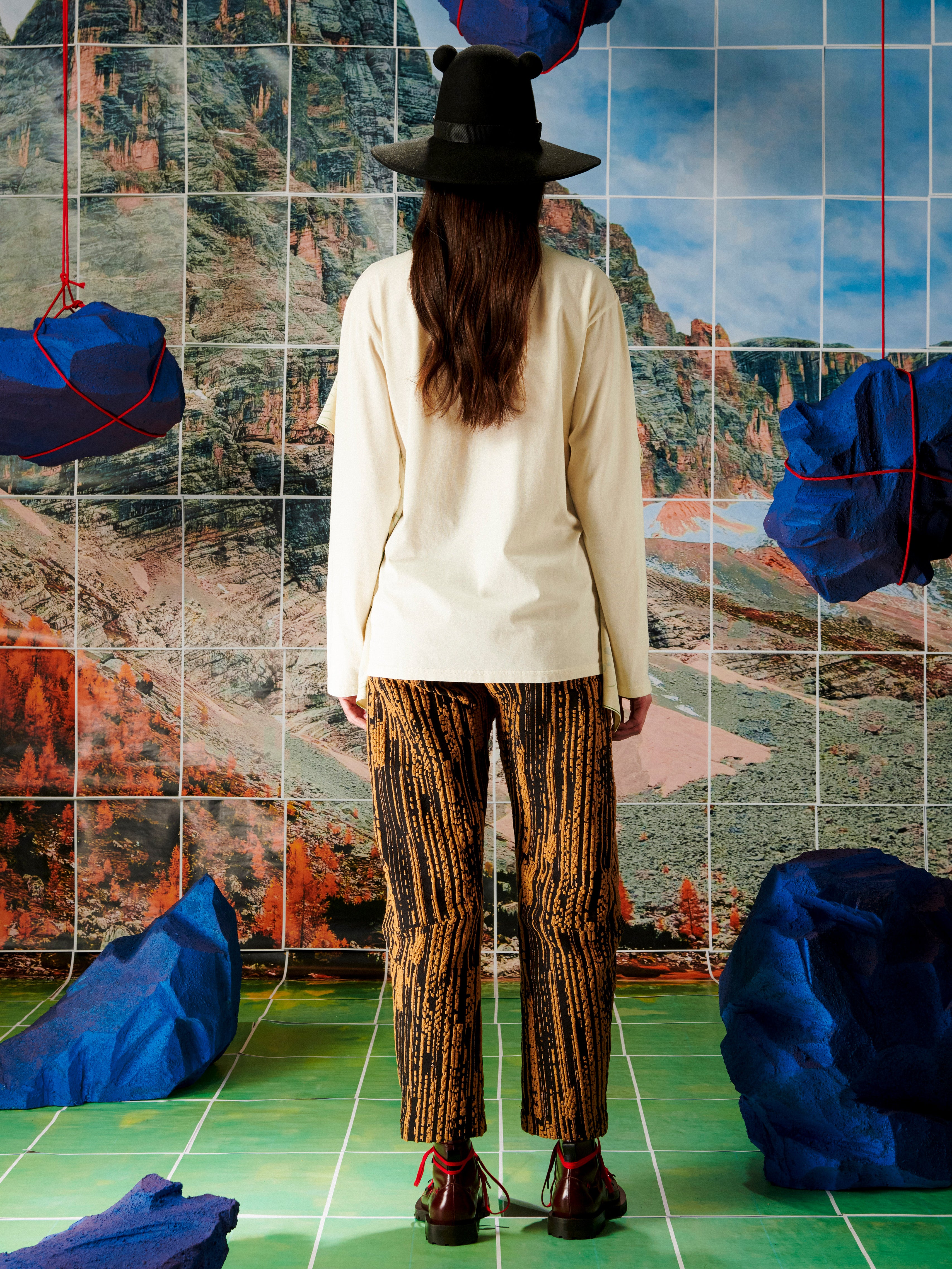 Person wearing a beige top and patterned pants standing in front of a tiled wall with a mountainous landscape design.