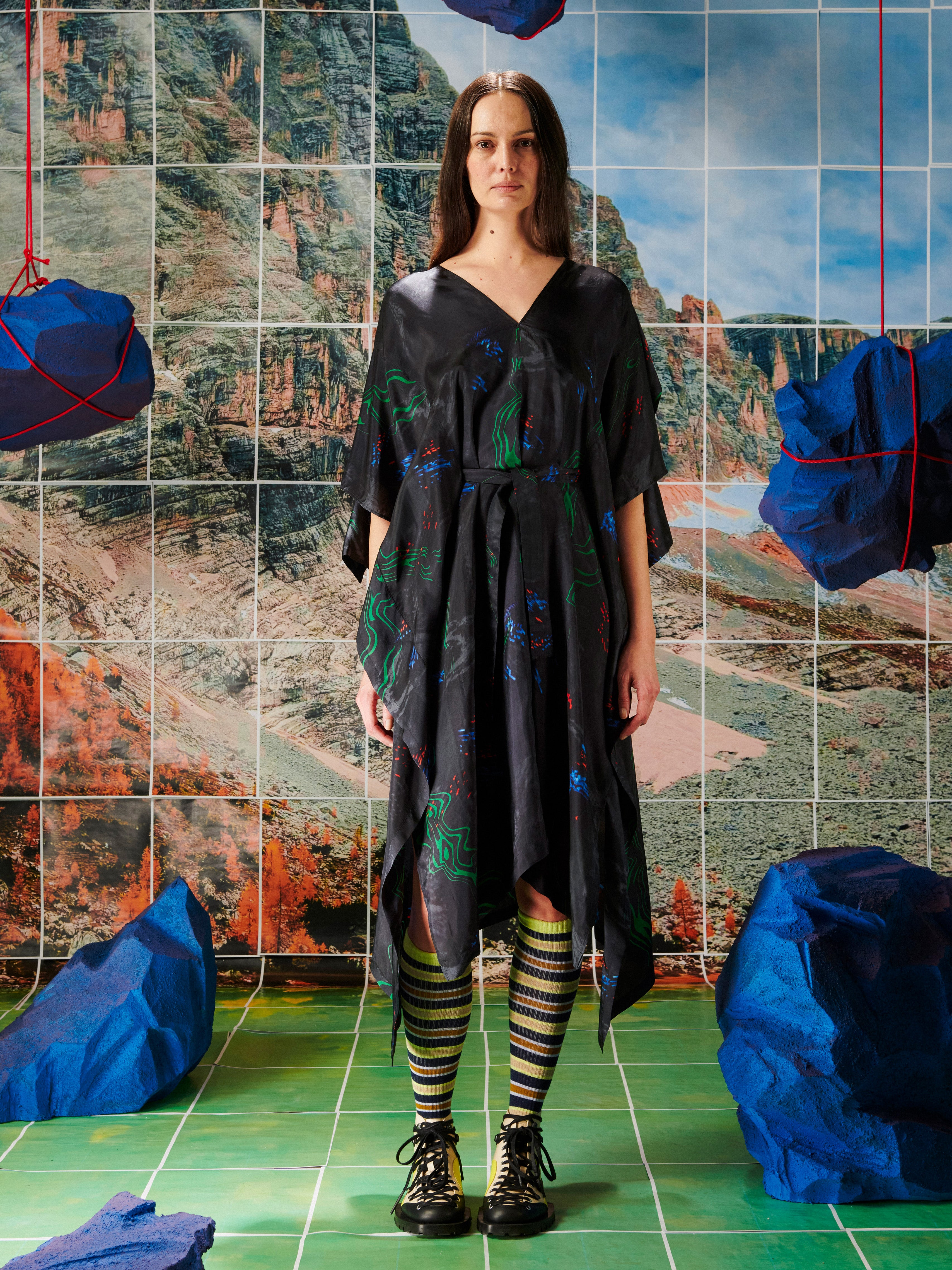Woman in a black dress with colorful patterns from Henrik Vibskov standing in front of a tiled wall with mountain design.