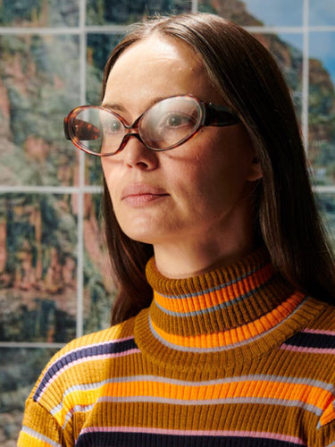 Person wearing glasses and a striped sweater in front of a colorful abstract painting