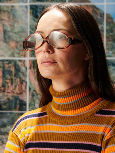 Person wearing large round glasses and a striped sweater in front of a colorful abstract painting.