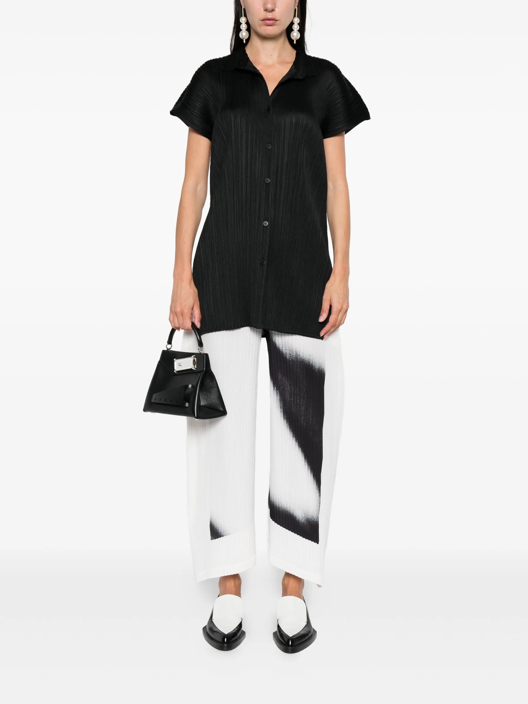 Person wearing a black pleated shirt and white pants with black accents, holding a black handbag on a white background.