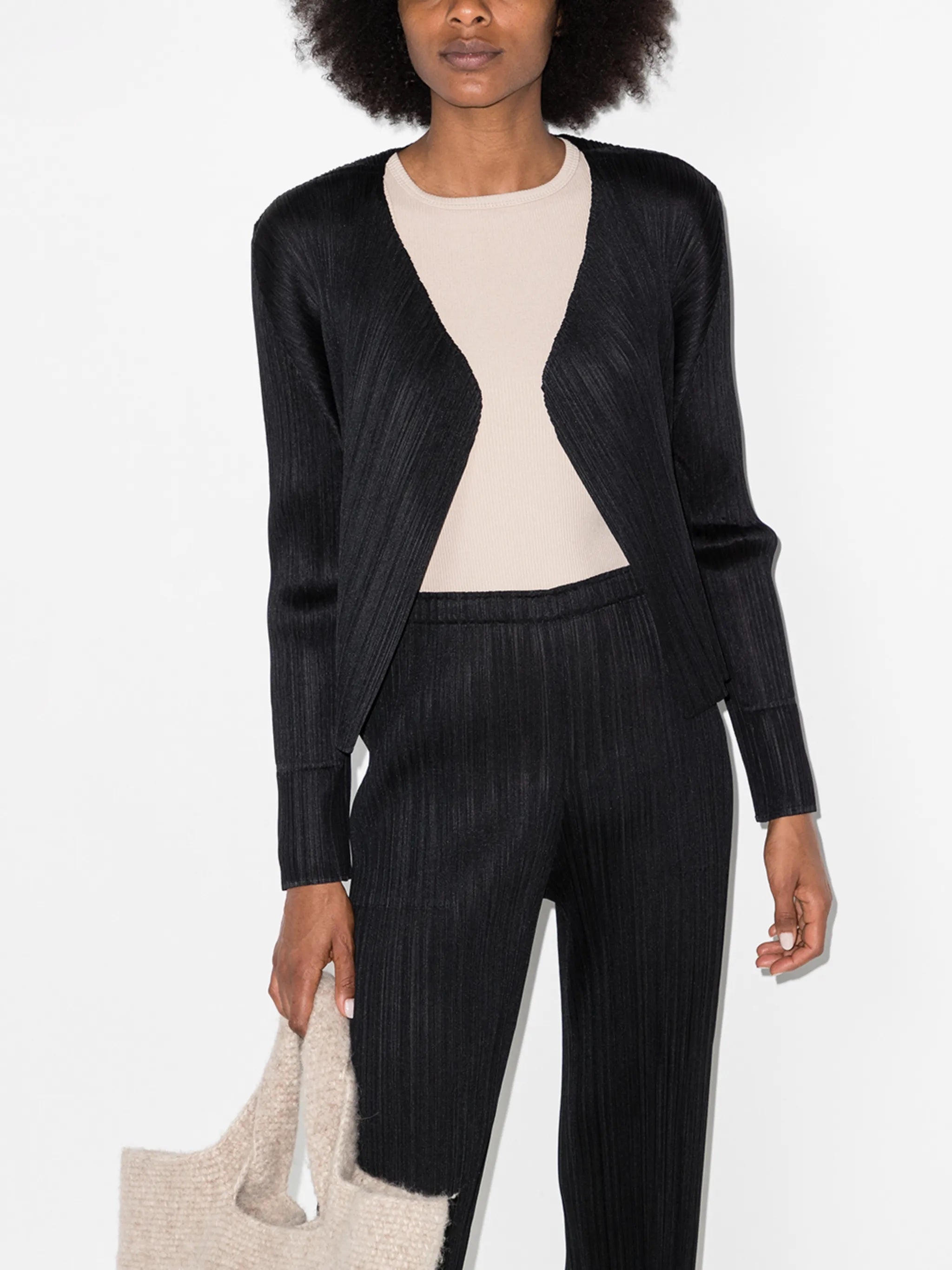 Woman wearing a black cardigan and black pleated pants holding a beige tote bag on a white background