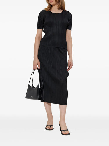 Woman wearing a black pleated top and a black pleated skirt holding a black handbag on a white background
