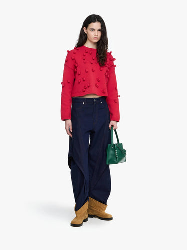 Woman wearing a red top with ruffled details, navy pants, and brown boots, holding a green handbag on a white background.