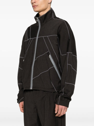 Close-up of model wearing the dark brown jacket with white geometric contrast stitching from Kiko Kostadinov on a white background