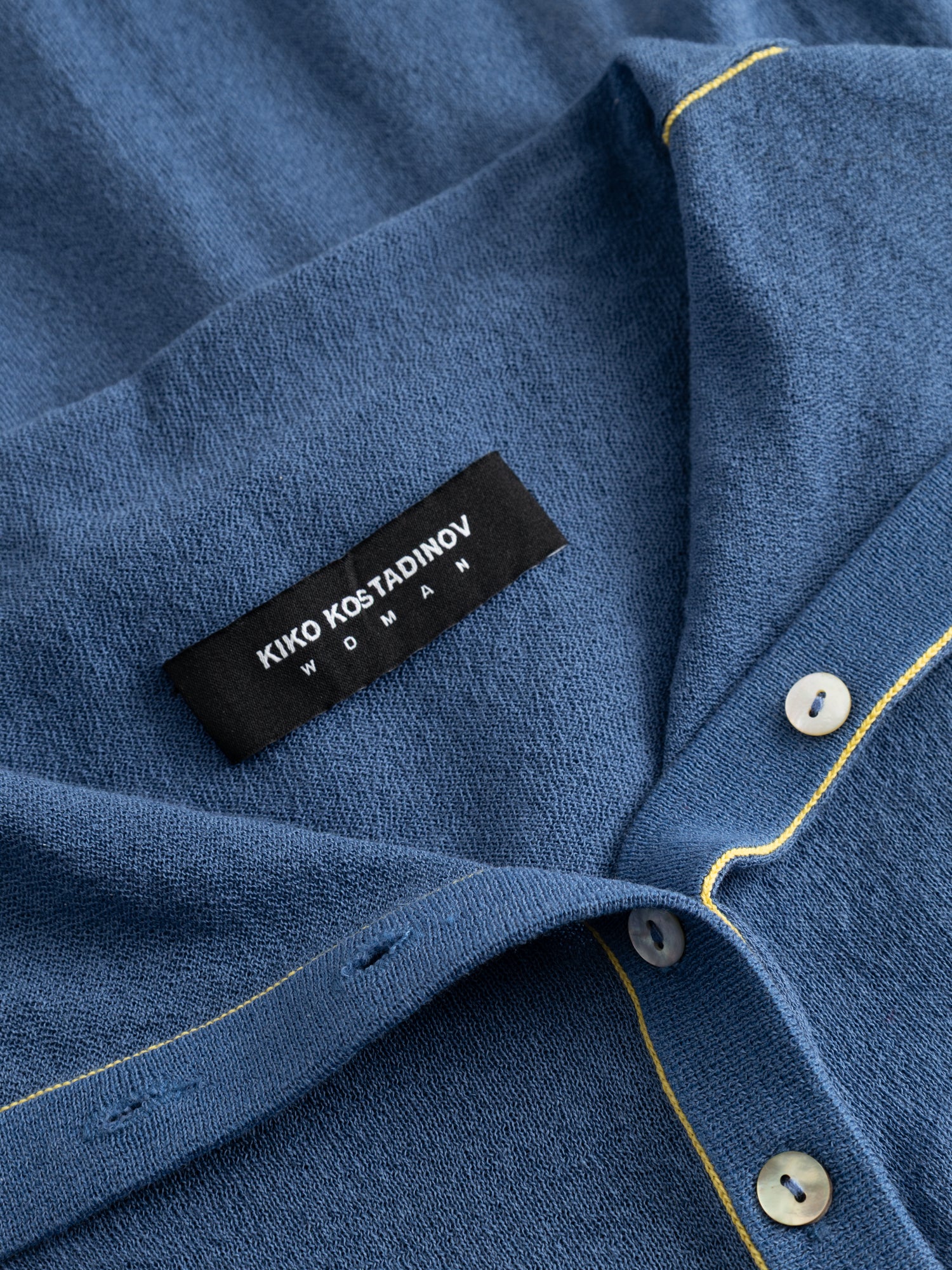 Kiko Kostadinov Woman Kreiner Knit Rollneck denim blue, close-up of the wide folded collar showing the woven brand label, yellow topstitched trim, and mother-of-pearl button detail at the neckline