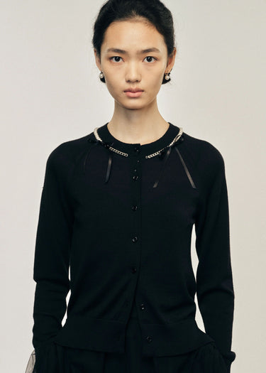 Person wearing a black cardigan with pearl and silver details with a plain background