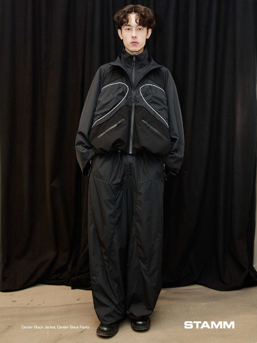 Model wearing the Stamm Exchange Center Black Jacket in Black with matching black trousers, showing the jacket’s high collar, curved piping and relaxed technical silhouette.