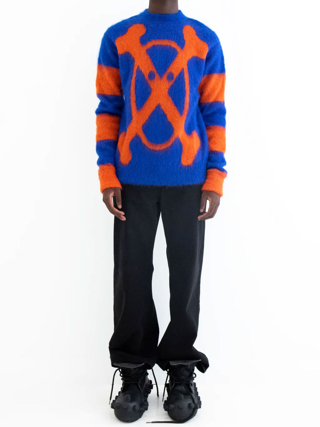 Model wearing Walter Van Beirendonck Crossed Bones Knit in Mazzarine Blue with orange graphic and sleeve stripes, paired with black trousers.