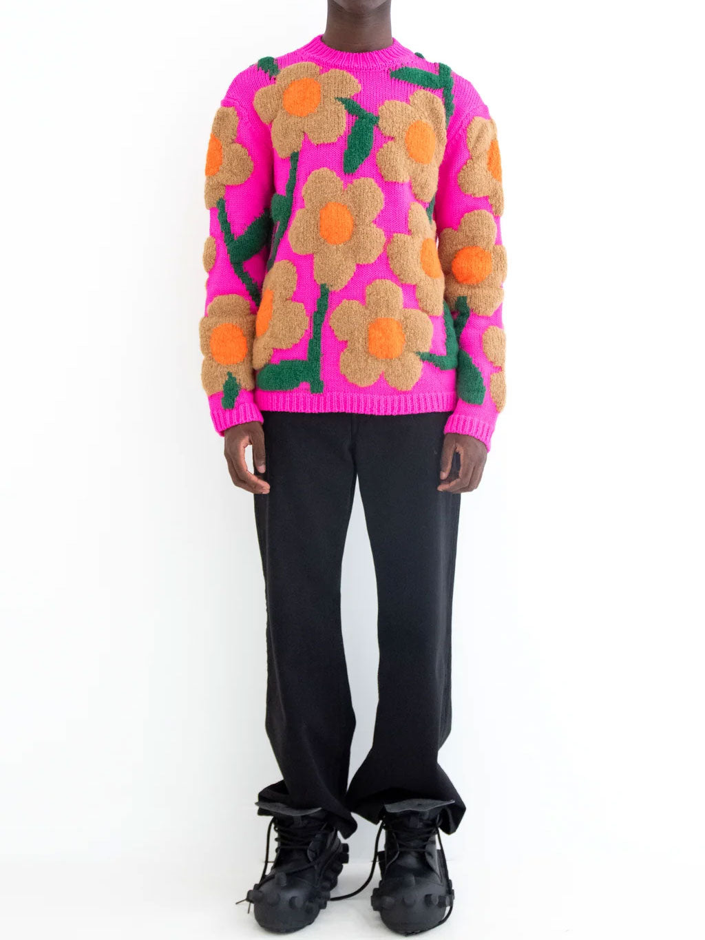 Model wearing Walter Van Beirendonck Flower Knit Sweater in pink with oversized floral motif, paired with black trousers and black shoes.
