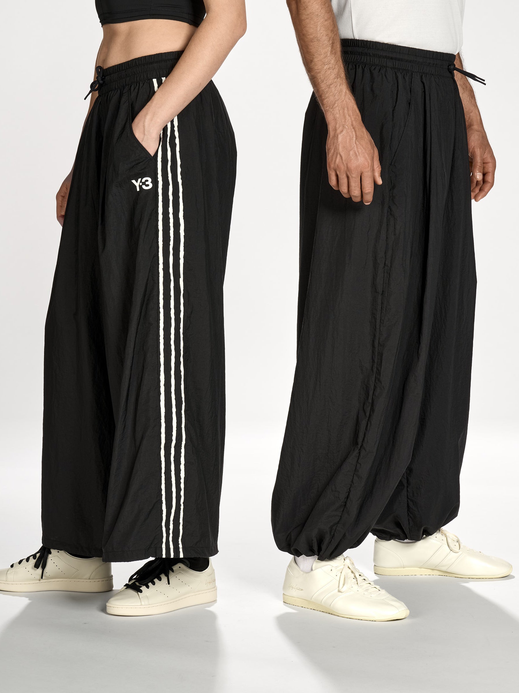 Side view of a female and male model wearing the Y-3 Raw Edge 3-Stripes Wide Tracksuit Bottoms in Black, emphasizing the loose, wide-leg fit, bungee-adjustable hems, and angled side pockets.