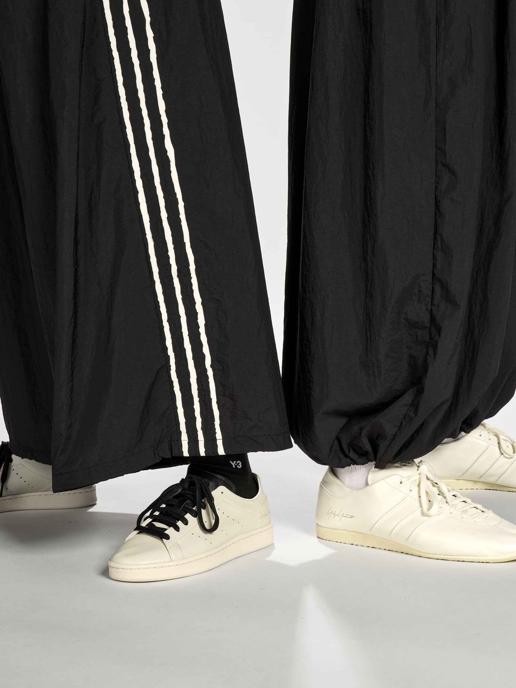 Close-up of a female and male model wearing the Y-3 Raw Edge 3-Stripes Wide Tracksuit Bottoms in Black, showing the texture of the fabric, raw-cut 3-Stripes detail, and satin heat-transfer Y-3 logo.