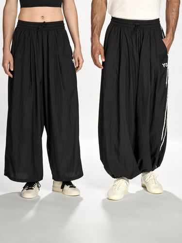 Front view of a female and male model wearing the Y-3 Raw Edge 3-Stripes Wide Tracksuit Bottoms in Black, showcasing the wide, relaxed silhouette, raw-cut 3-Stripes at the right leg, and satin Y-3 logo at the left front.